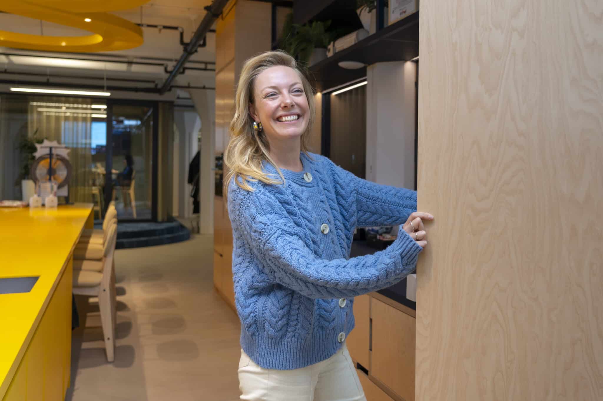 A smiling woman wearing a blue knit sweater stands indoors, holding the edge of a light wooden sliding panel in a modern office space with yellow tables, shelving, and soft lighting in the background.